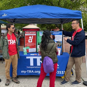 Two men in red shirts standing by outreach table speak to woman whose back is to the camera